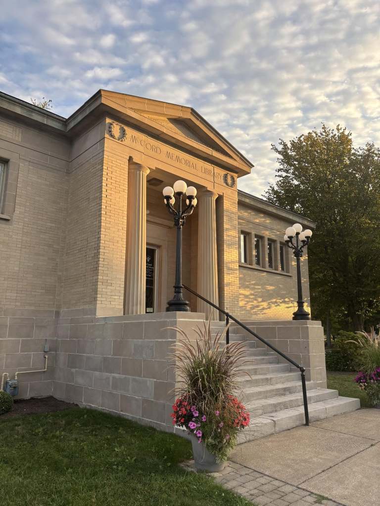 The library from the front with speckled clouds in the blue sky. The library is at an angle and the planters are in the front of the steps.